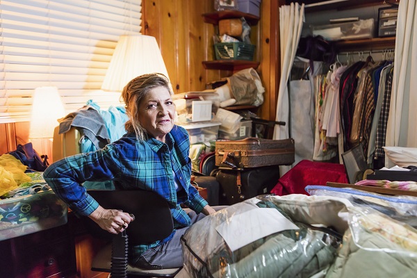 A senior woman in her 60s at home, sitting in a messy, cluttered room, looking at the camera with a serious expression.