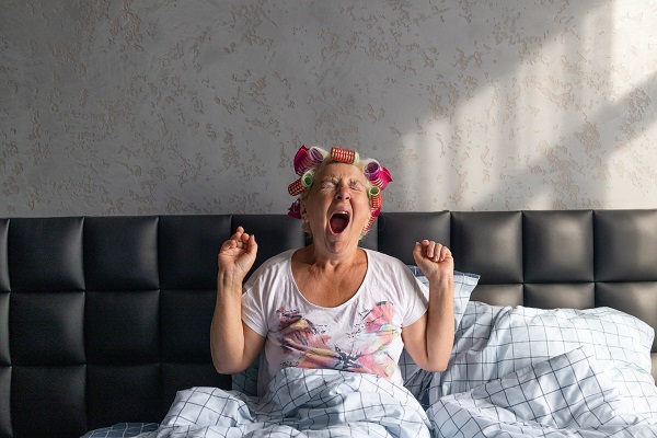 Picture of a tired middle aged woman in curlers wake up yawns sitting on bed at home bedroom.