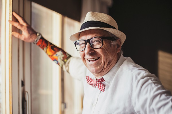 Older adult man with sleve tattoo standing by window smiling and looking at camera