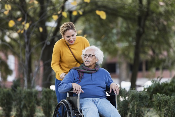 A senior Hispanic man in his 80s sitting in a wheelchair, taking a walk in the park with his adult daughter, a mid adult woman in her 30s.